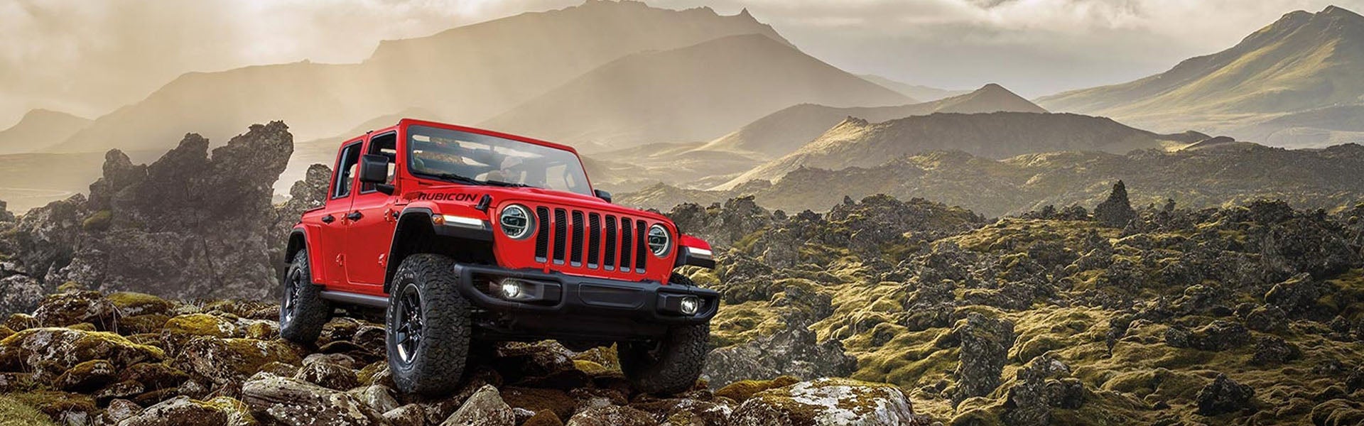 A pre-owned bright red Jeep Wrangler Rubicon parked on a rocky off-road terrain with mountains in the background.