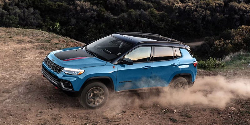 A blue Jeep Compass Trailhawk driving off-road on a dirt trail, kicking up a plume of dust.