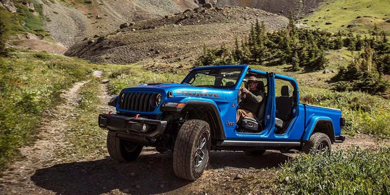 A blue Jeep Gladiator Mojave pickup truck, doors removed, driven by a man on a dirt trail in a mountain valley.