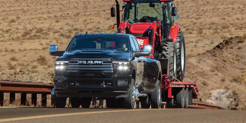 A black Ram dually pickup truck tows a red tractor on a flatbed trailer down a desert highway.