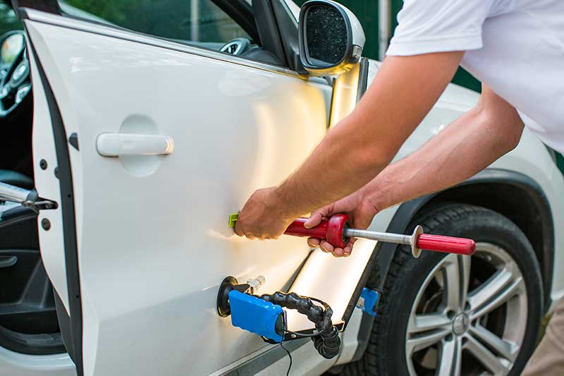A person uses specialized tools to remove a dent from the white door panel of a car.