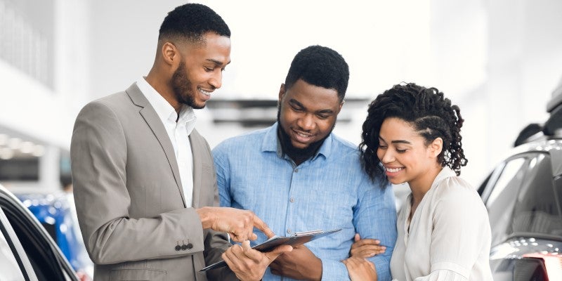 Car salesman pointing at a contract on a clipboard for a smiling couple in a busy dealership.