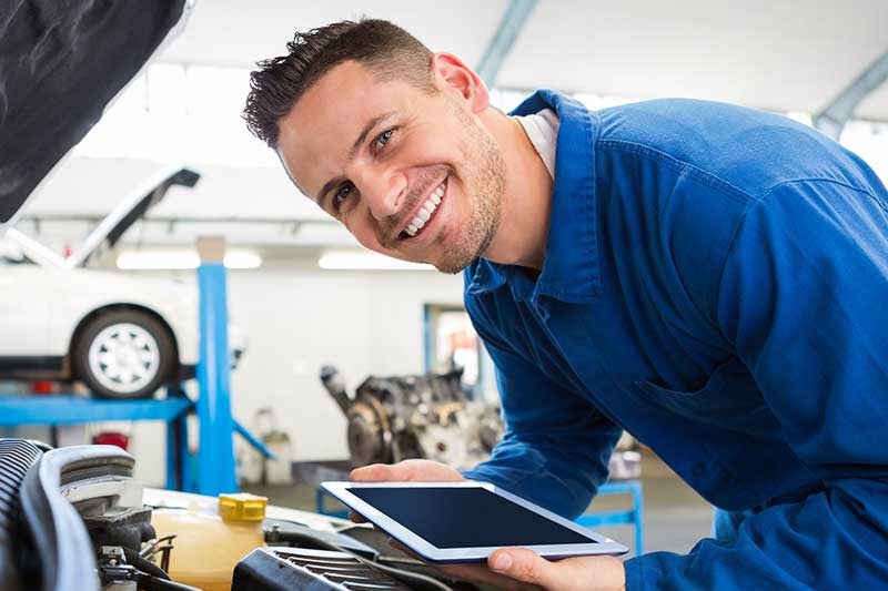 Smiling mechanic in blue coveralls holding a tablet while leaning over a raised car's engine in a garage.